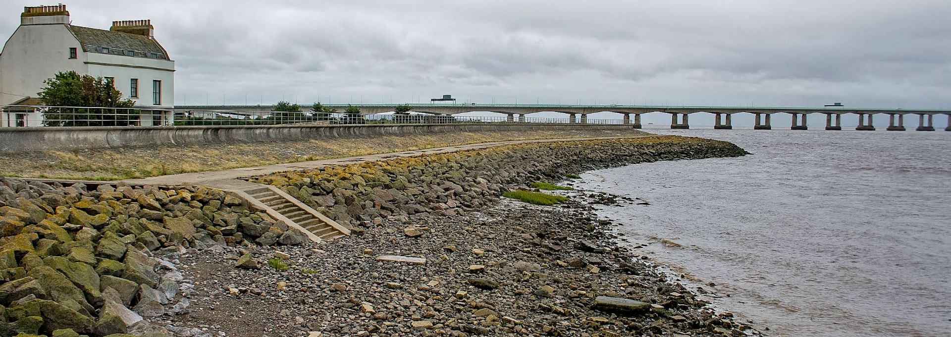 View of the rocky Severn Beach shoreline with a bridge and lighthouse in the background.