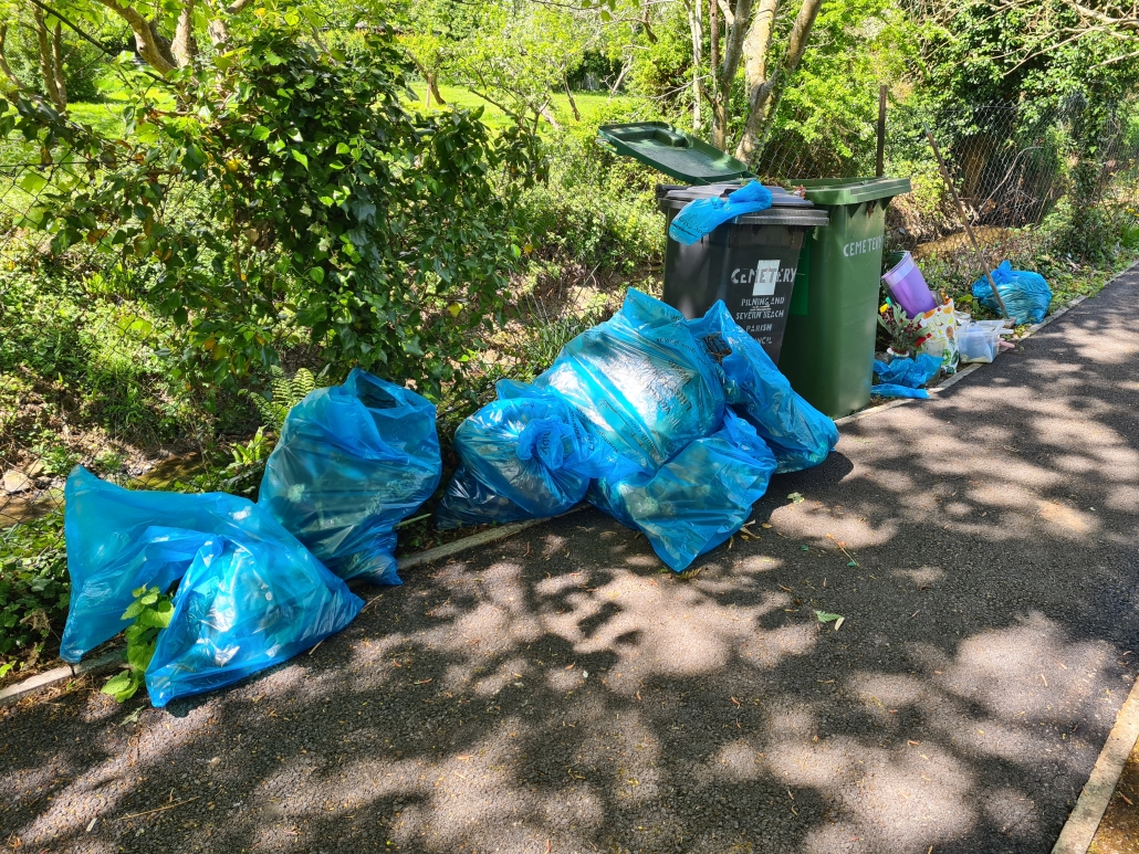 Several blue bin bags piled beside overflowing green bins on a sunny path near trees and bushes.