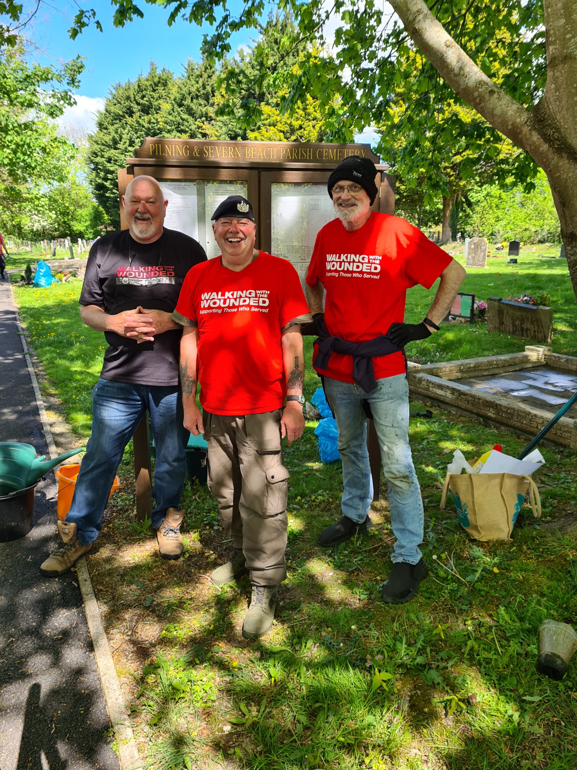 Three smiling men in "Walking With The Wounded" shirts stand together in a sunny cemetery.