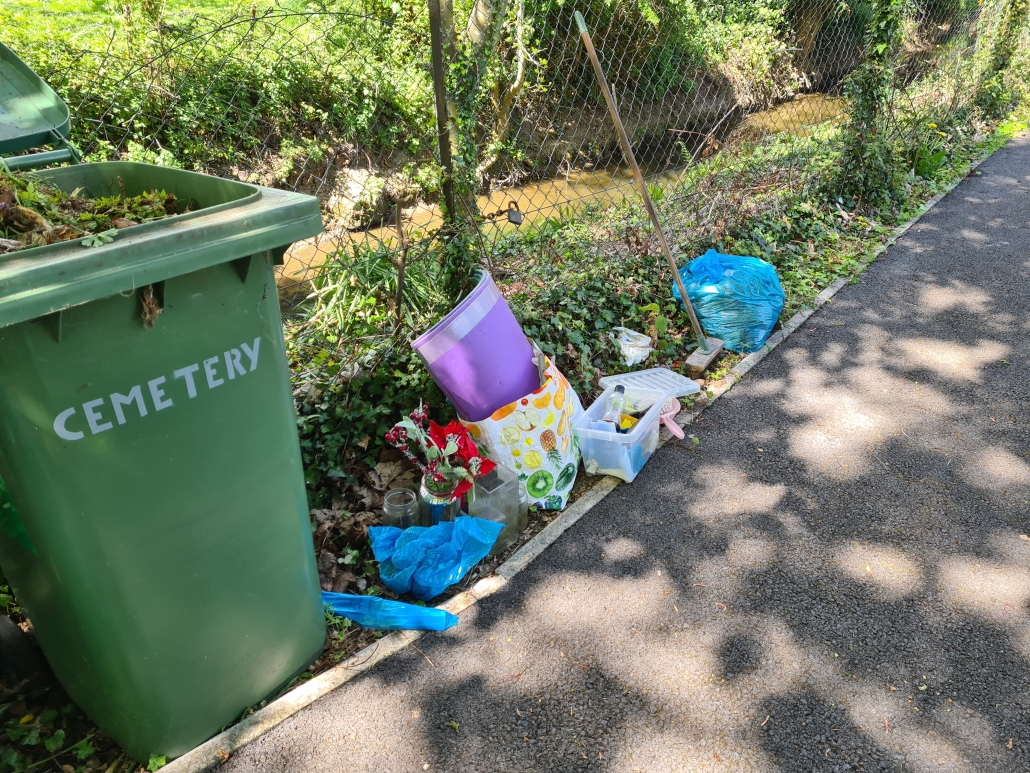 A cemetery bin and discarded items, including flowers and plastic bags, sit beside a sunlit path and fence.